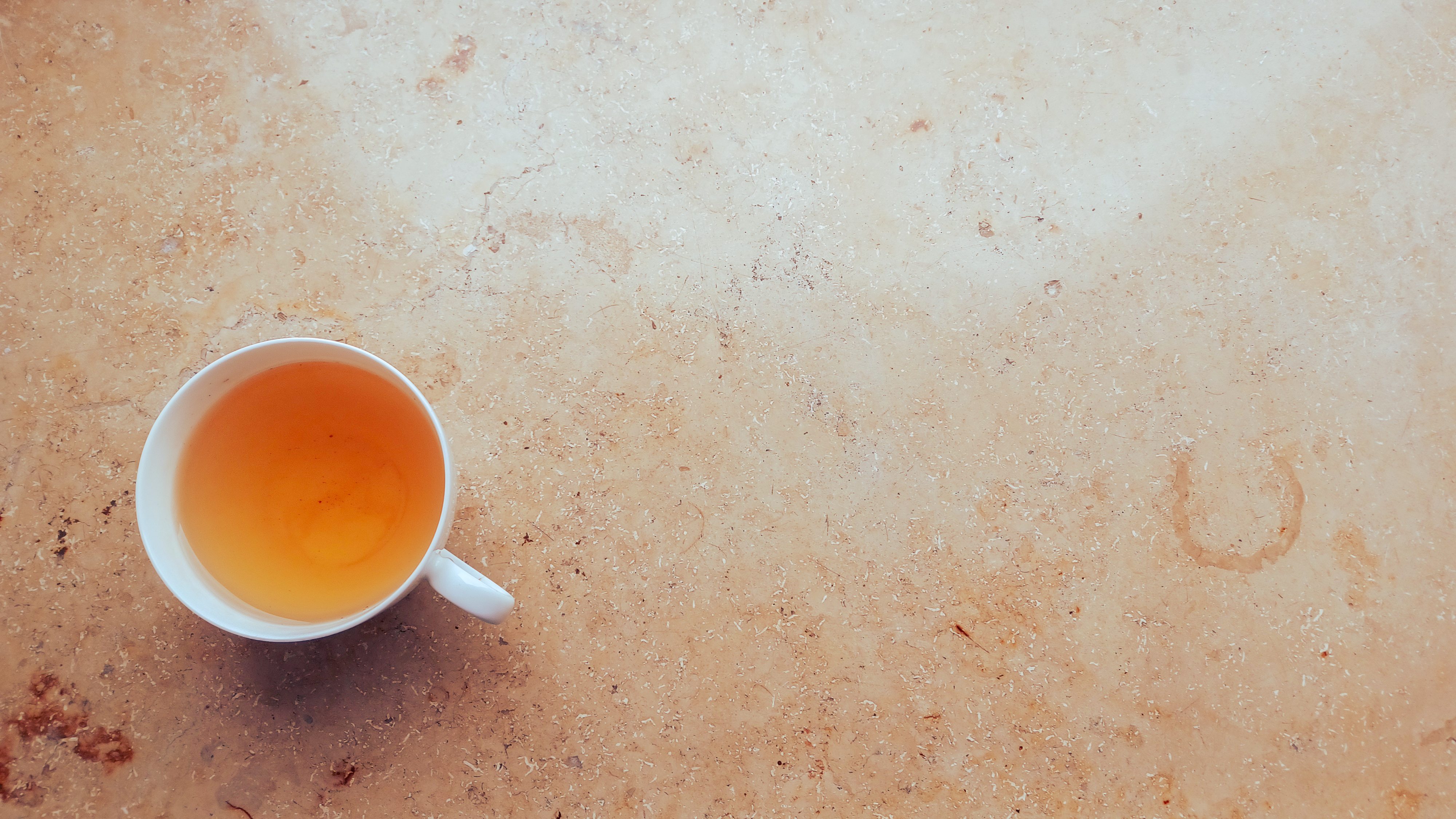 Foto einer weissen Teetasse aus Porzellan mit goldgelbem Tee. Die Tasse steht auf einer beige-braunen Natursteinplatte. 